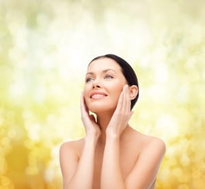 A woman with glowing skin smiling and touching her face on a yellow and white light background