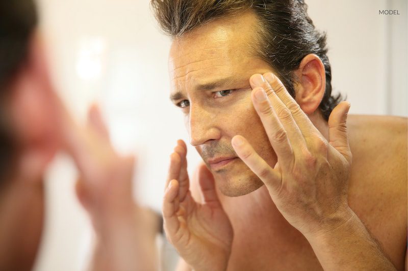 middle-aged man looking in mirror, pulling skin back near his eye, determining skin laxity