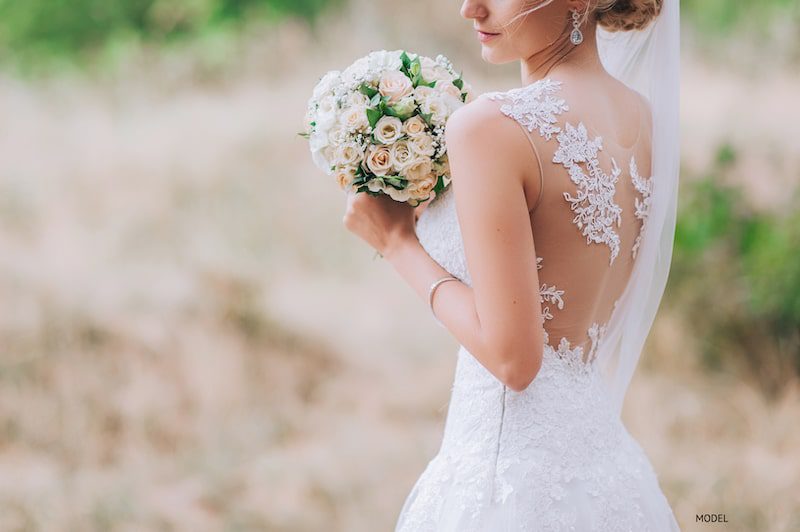 Image of a woman's back, wearing a lacy, sleeveless wedding dress, holding a bouquet of flowers.