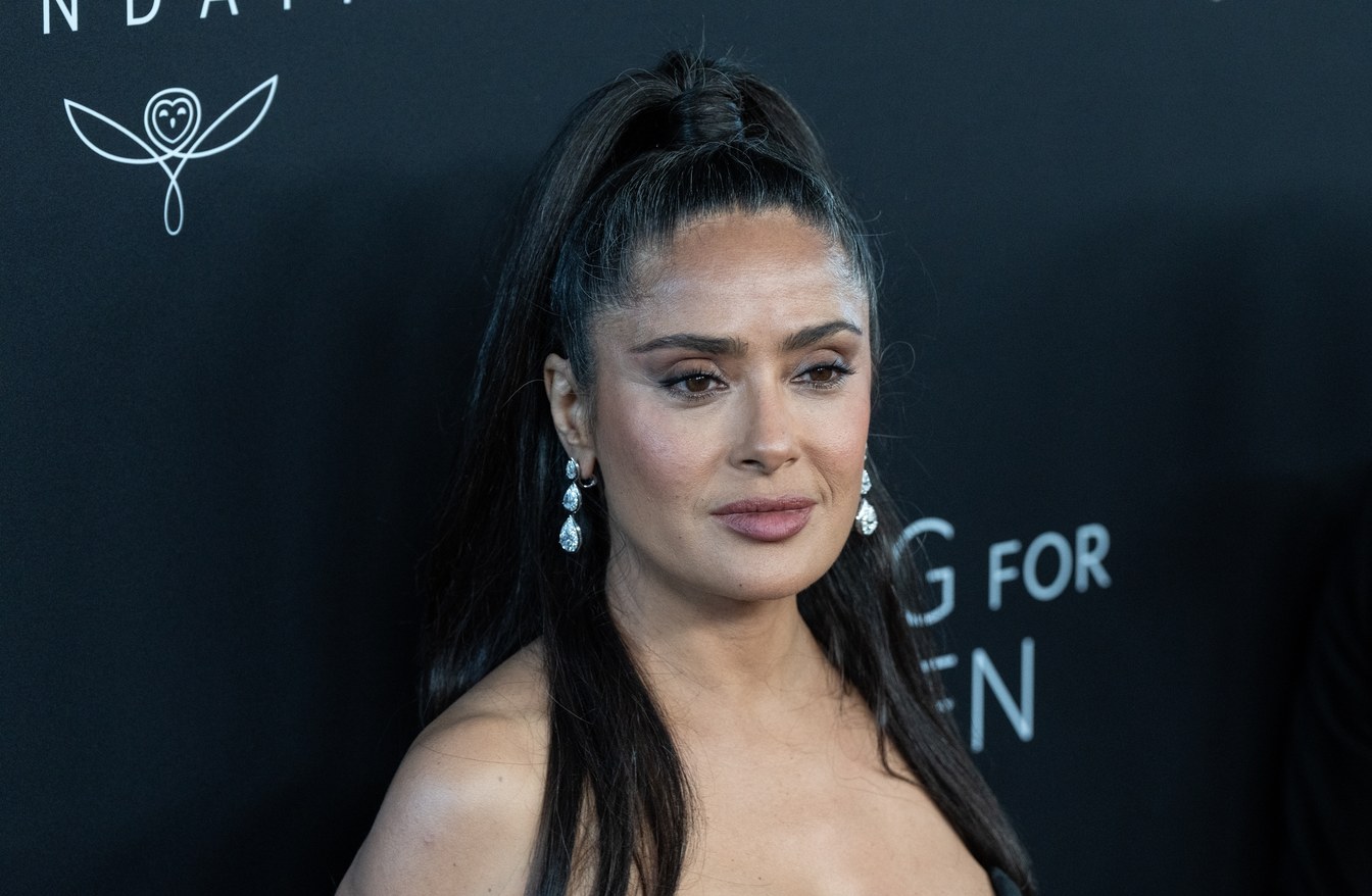 Salma Hayek on the red carpet in September 2025, long dark hair in a high half ponytail, drop diamond earrings, photographed at a three-quarter angle. Commentary by Frank Agullo, MD, FACS.